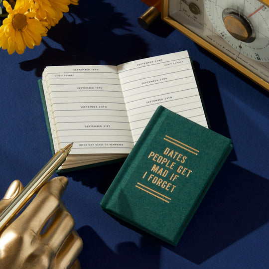 A green, hard-bound Brass Monkey Dates People Get Mad If I Forget Tiny Diary with gilded-edge pages rests on a blue surface beside an open planner, a gold hand-shaped pen holder, a pen, yellow flowers, and part of a vintage scale.