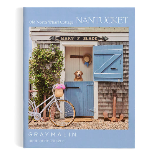 The Gray Malin Nantucket 1000 Piece Book Box Puzzle showcases Nantucket photography—a cottage with a dog at the blue door, flowers, a white bicycle, and the Mary F. Slade sign. Brand: Gray Malin.