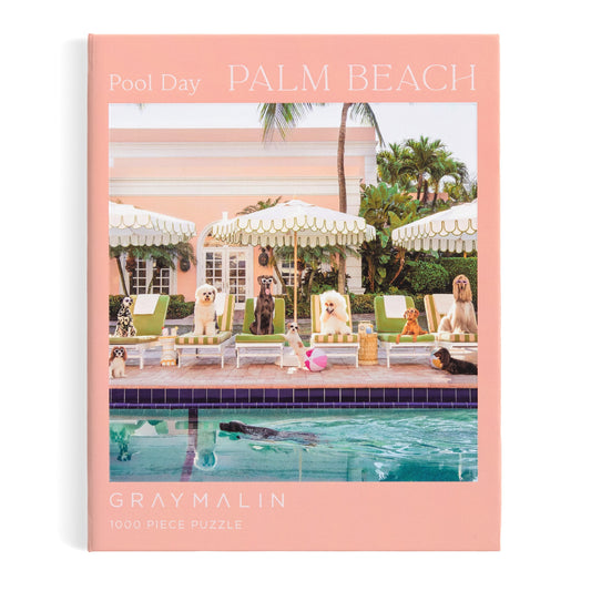The Gray Malin Pool Day Palm Beach 1000 Piece Book Box Puzzle by Gray Malin shows dogs relaxing by a pool with The Colony Hotel’s pink facade and palm trees, and features Pool Day Palm Beach and Gray Malin in white on the cover.