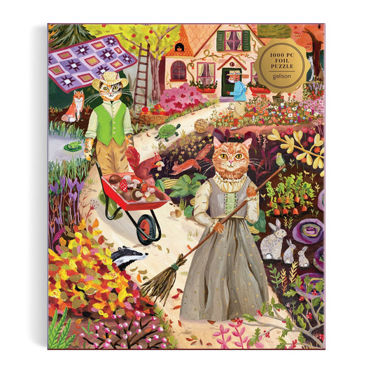 The Anja Riebensahm Cottage Cats 1000 Piece Foil Puzzle features whimsical, illustrated cats in period clothing gardening among flowers near a cottage—one cat pushes a wheelbarrow, another sweeps the path.