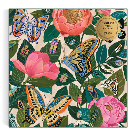 The Between the Blooms 1000 Piece Foil Puzzle by Christine De Carvalho features butterflies, beetles, and moths among pink flowers and green leaves, with a gold label that reads “1000 piece foil puzzle.”.