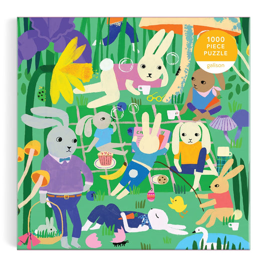 The Bunny Daydream 1000 Piece Puzzle by Anne Bentley features rabbits picnicking, playing games, blowing bubbles, and relaxing among large flowers in a grassy field. The puzzle box displays the piece count in the top right corner.