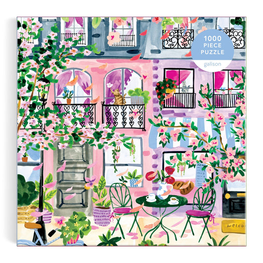 Cherry Lane Blossoms 1000 Piece Puzzle by Sabina Fenn features a vibrant pink building with balconies, blooming flowers, lush greenery, and a café table with green chairs on a patio—perfect for springtime street scene lovers.