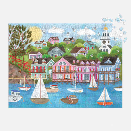 The Joy Laforme Harbor by the Sea 1000 Piece Puzzle by Joy Laforme features sailboats in a Nantucket harbor, colorful hillside houses, trees, and a white church under a blue sky, with several puzzle pieces missing.