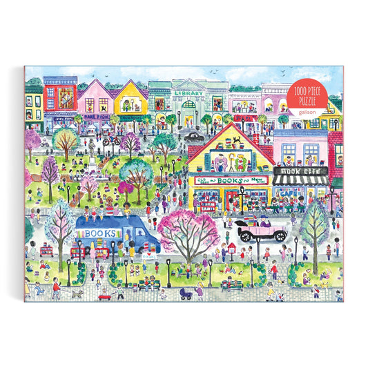 The Michael Storrings Book Town 1000 Piece Puzzle by Michael Storrings features a vibrant town scene with colorful buildings, a park, people, libraries, bookstores, a bookshop van, and an old-fashioned car in a lively atmosphere.