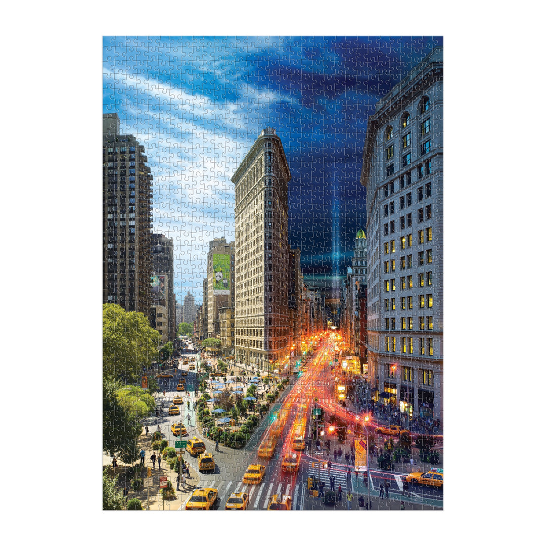 The Stephen Wilkes Day To Night Flatiron 1000 Piece Puzzle by Stephen Wilkes features a New York Cityscape with tall buildings, yellow taxis, busy streets, and the iconic Flatiron Building transitioning from day to night.