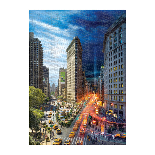 The Stephen Wilkes Day To Night Flatiron 1000 Piece Puzzle by Stephen Wilkes features a New York Cityscape with tall buildings, yellow taxis, busy streets, and the iconic Flatiron Building transitioning from day to night.