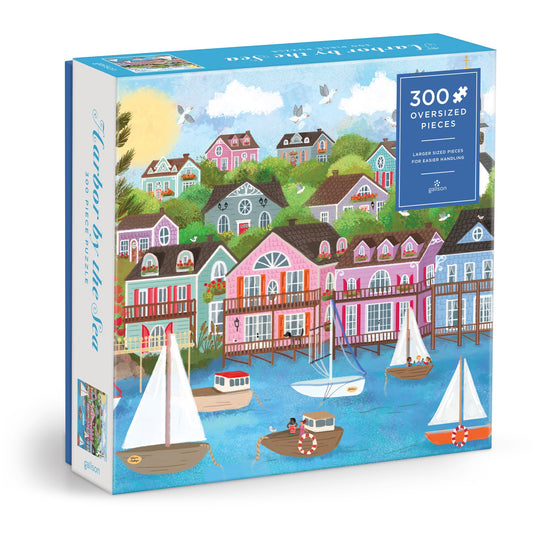 The Joy Laforme Harbor by the Sea 300 Piece Puzzle features vibrant seaside houses, boats, trees, and birds in a colorful design by Joy Laforme. The box highlights 300 oversized pieces for a fun and vivid puzzle experience.