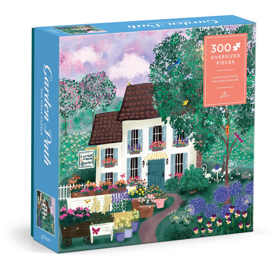 The Joy Laforme Garden Path 300 Piece Puzzle by Joy Laforme features a white house with a brown roof, surrounded by vibrant gardens and flowers, all beneath a pink and blue sky.