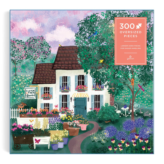 The Joy Laforme Garden Path 300 Piece Puzzle by Joy Laforme features a charming house with a brown roof, vibrant flowers, a garden path, and trees under a whimsical pink and purple sky. A sign reads “Clover & Fern Garden.”.