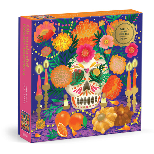 The Marisol Ortega Ofrenda del D’a de Muertos 500 Piece Foil Puzzle features a sugar skull, bright flowers, fruit, pastries, and candles on a purple background with gold foil accents celebrating Mexican culture.