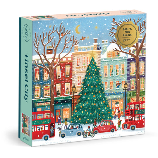 The Laura Watkins Tinsel City 500 Piece Foil Puzzle features a festive urban holiday scene with colorful buildings, a big decorated Christmas tree, red London-style buses, cars, and snowy streets.