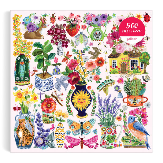 The Buzzy Bouquets 500 Piece Puzzle by Jennifer O Lewis/August Wren features a whimsical, nature-inspired illustration with painted flowers, butterflies, birds, a cottage, and a rabbit amid vibrant plants.