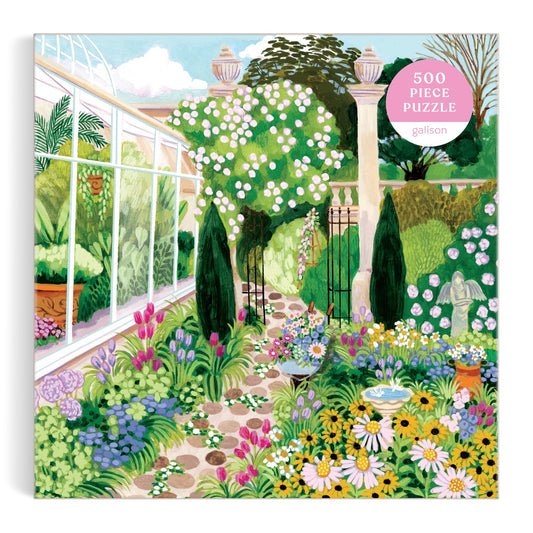 The Evergreen Gardens 500 Piece Puzzle by Joanna Clay features an illustration of a lush garden with colorful flowers, bushes, a stone path, fountain, and greenhouse on the box.
