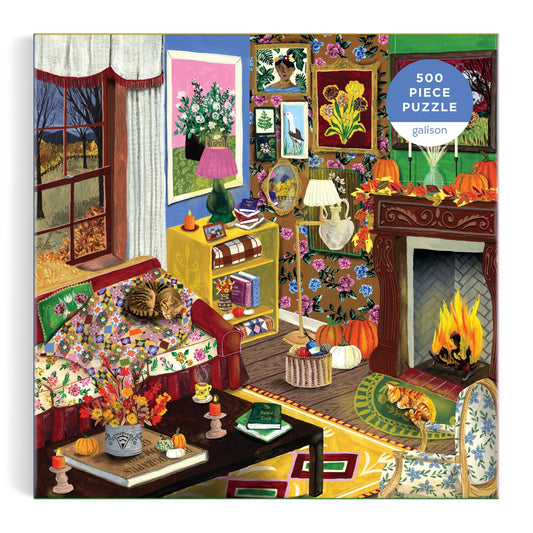 Enjoy a cozy living room scene with a cat lounging on a floral couch, a fireplace aglow with fire, autumn decorations, books, and art adorning the walls—plus a window framing a beautiful fall landscape. This charming image is featured on Abby Jacobs' Fall is in the Air 500 Piece Puzzle—a perfect rainy day activity, beautifully packaged in an eye-catching box.