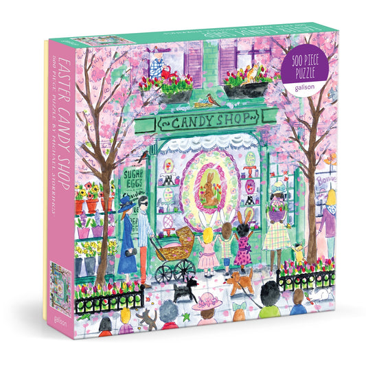 The Michael Storrings Easter Candy Shop 500 Piece Puzzle features a vibrant scene with children, bunnies, Easter eggs, and spring décor under blooming pink trees. The box displays “Easter Candy Shop, 500 Piece Puzzle.”.