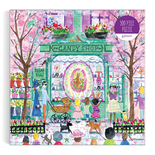 The Michael Storrings Easter Candy Shop 500 Piece Puzzle by Michael Storrings features a lively candy shop with pink blossoming trees, Easter eggs in the window, and a festive crowd outside in a vibrant, colorful scene.