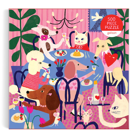 The Paws for Love 500 Piece Puzzle by Anne Bentley features a colorful illustration of six dogs and one cat toasting drinks around a blue table, with pink striped wallpaper and plants in the background.