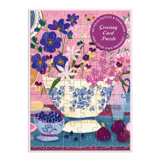 The Joy Laforme Art of the Table Greeting Card Puzzle by Joy Laforme shows a vase with blue flowers, a berry bowl, and figs on a table set against a pink and purple background. A round “Greeting Card Puzzle” label is included.