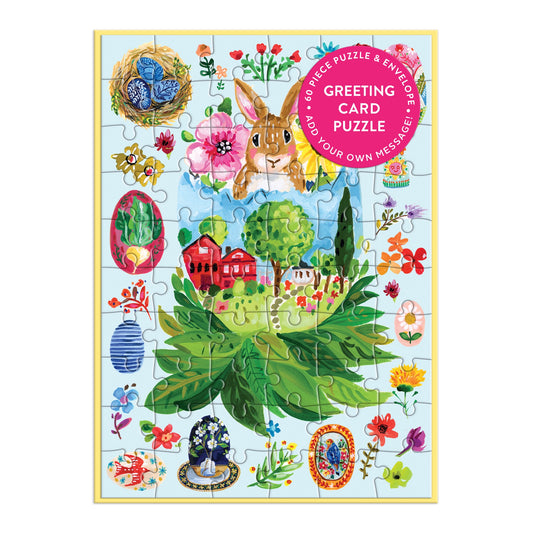 The Artisanal Eggs Greeting Card Puzzle by Jennifer O Lewis /August Wren features a rabbit, red house, green trees, flowers, and eggs. A pink sticker says Greeting Card Puzzle—the perfect Easter wish to send!.