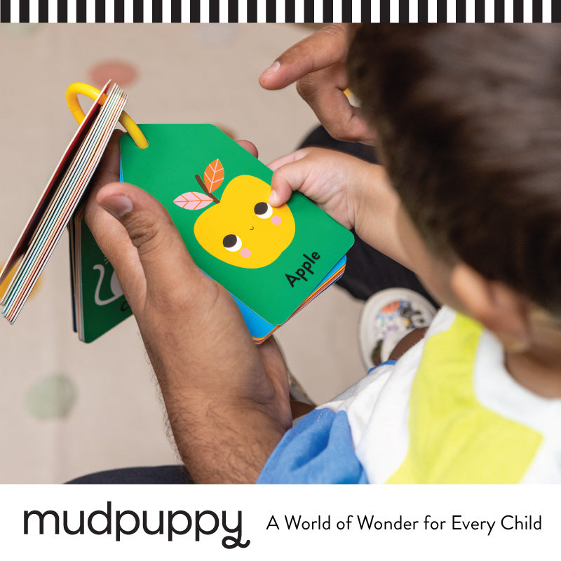 Adult and child hands holding a colorful board book page featuring a smiling yellow apple illustration with the word Apple written below it. The book is shaped like tags and attached by a yellow ring.