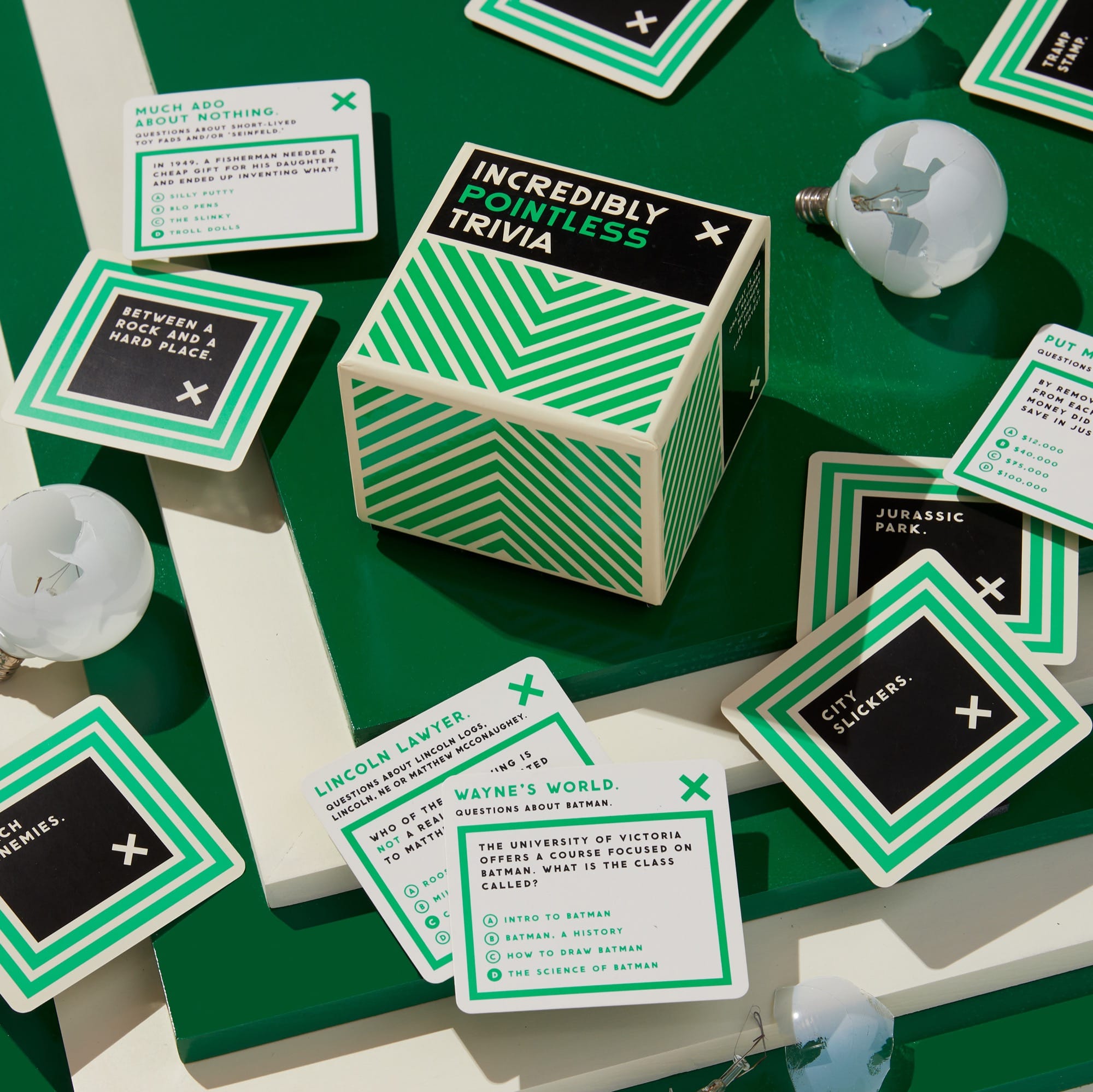 A box labeled Incredibly Pointless Trivia sits on a green surface, surrounded by trivia cards and broken light bulbs. The cards feature various trivia questions and answers.