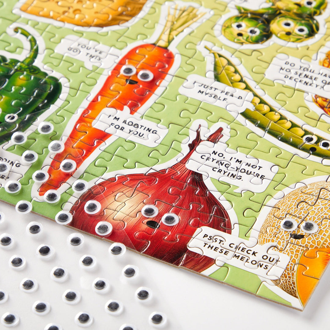 The Brass Monkey Googly Food 1000 Piece Puzzle features illustrated vegetables with playful speech bubbles and googly eyes, plus a sheet of unused googly eyes in the corner for extra fun in this quirky food-themed collage.