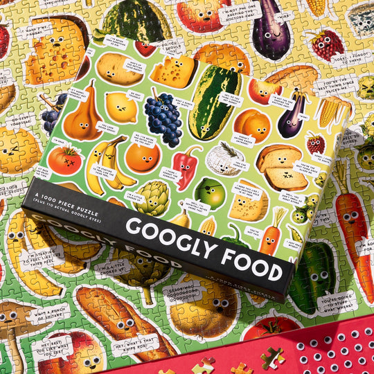 The Brass Monkey Googly Food 1000 Piece Puzzle, featuring a fun collage of fruits and veggies with googly eyes, is shown atop a finished puzzle with a red tray at the bottom.