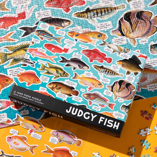 The Brass Monkey Judgy Fish 1000 Piece Puzzle features a colorful box with humorous fish illustrations, spot UV accents, and eco-friendly pieces scattered beside a partially completed fish-themed puzzle.