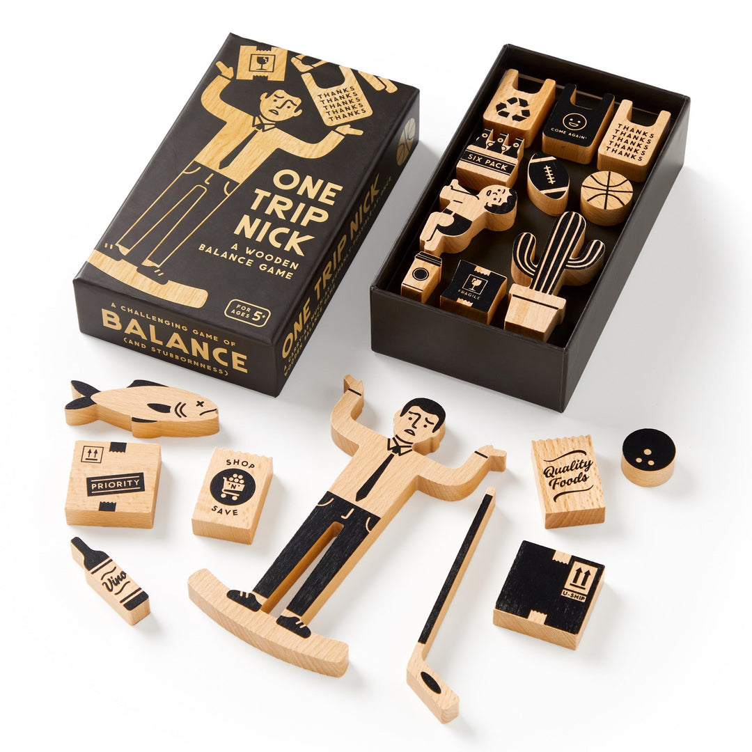 The One Trip Nick Wooden Balance Game by Brass Monkey features a box and unique black and natural wood grocery-themed pieces, like a fish and plant. Some pieces are displayed outside the box.