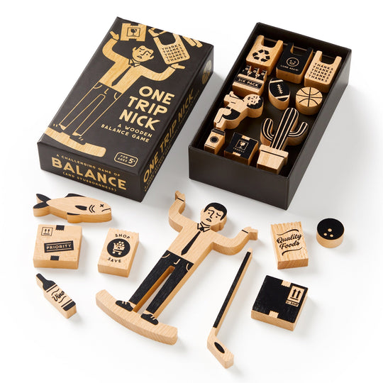 The One Trip Nick Wooden Balance Game by Brass Monkey features a box and unique black and natural wood grocery-themed pieces, like a fish and plant. Some pieces are displayed outside the box.