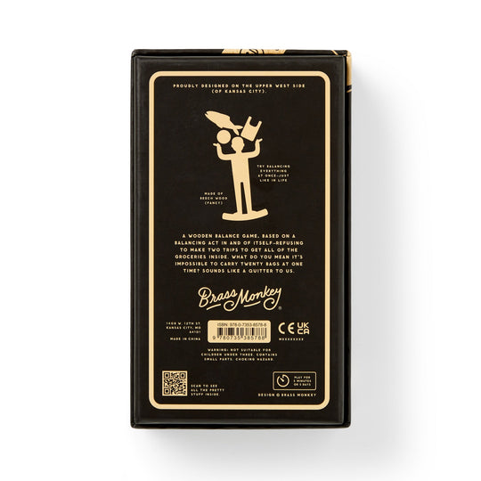 The back of a black Brass Monkey game box displays a gold illustration of balancing objects, descriptive text about the One Trip Nick Wooden Balance Game with grocery-themed items, plus branding, barcode, and certification icons at the bottom.
