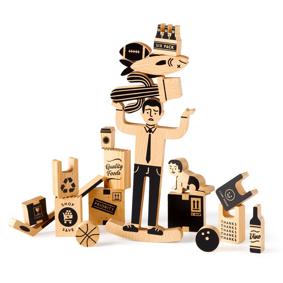 The One Trip Nick Wooden Balance Game by Brass Monkey features a central man holding items like a football, fish, and six-pack, surrounded by wood pieces shaped as food, a baby, basketball, and grocery-themed objects.
