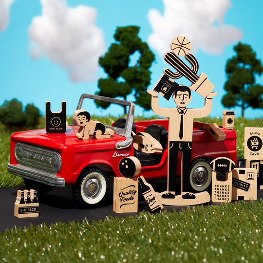 The One Trip Nick Wooden Balance Game by Brass Monkey features wood game pieces shaped like a family and groceries, set beside a red toy convertible on grass under a blue sky with clouds.