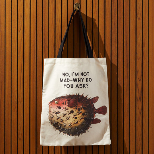 The Brass Monkey Judgy Fish Canvas Tote, a white humorous bag with black handles, hangs on a wooden slatted wall. It showcases vintage pufferfish art and the phrase NO, IM NOT MAD—WHY DO YOU ASK? above the image.