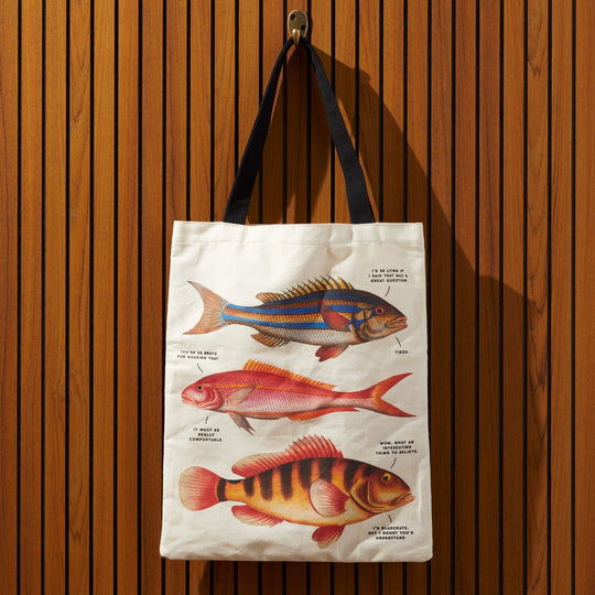 The Judgy Fish Canvas Tote by Brass Monkey features three vintage-inspired, illustrated fish with witty captions on a white bag with black handles, shown hanging vertically against a wooden wall.