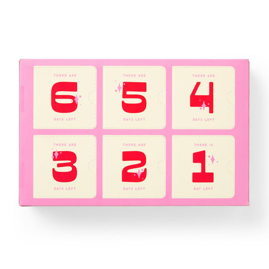 The Brass Monkey Googly & Bright Holiday Ornament Countdown Calendar features six numbered pink compartments, each with bold red digits on cream, perfect for building holiday excitement or storing small ornaments.