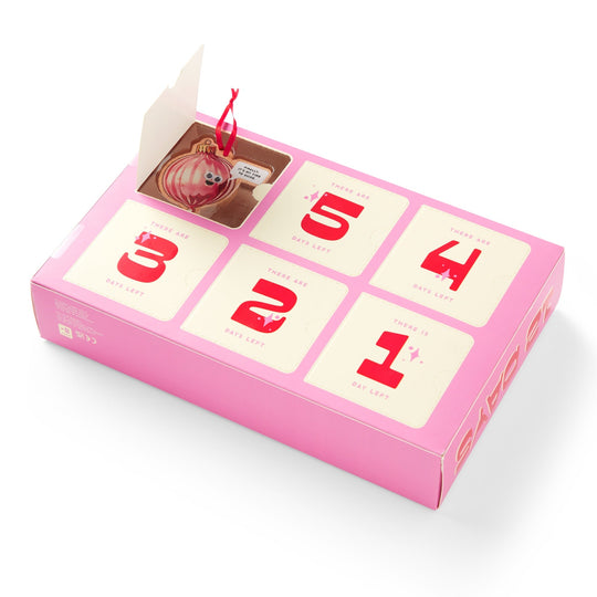 Brass Monkey’s Googly & Bright Holiday Ornament Countdown Calendar features 6 pink numbered flaps counting down the days, with playful ornaments—like a Piglet with a red string and googly eyes—hidden behind each flap.