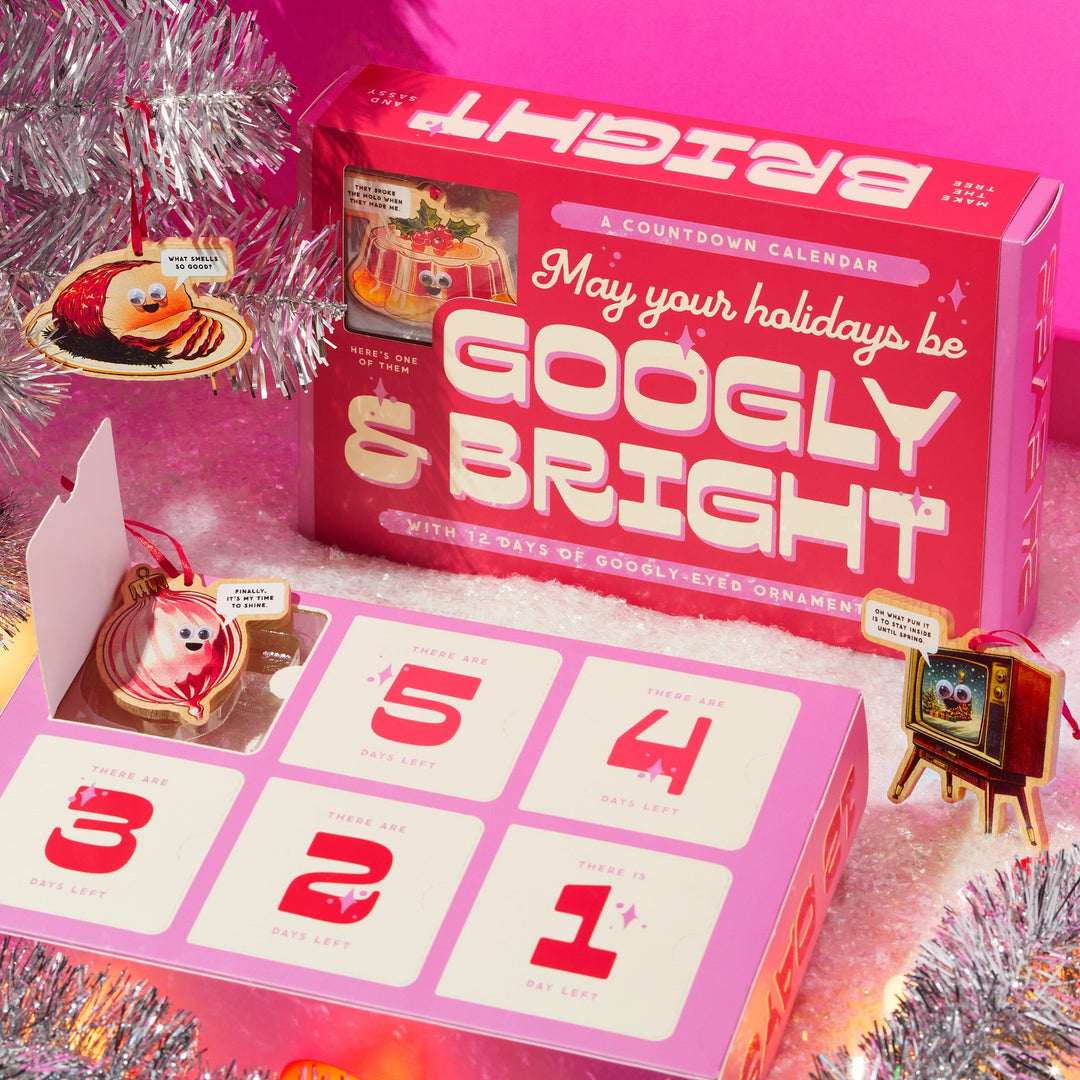 A Googly & Bright Holiday Ornament Countdown Calendar by Brass Monkey sits open, displaying a googly-eyed ornament inside amid silver tinsel and colorful holiday decor.