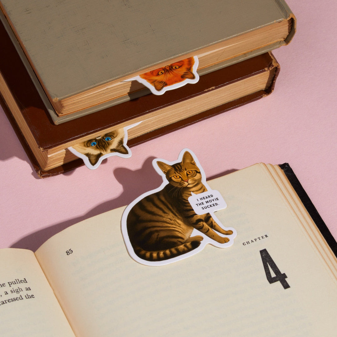 The Brass Monkey Moody Cats Magnetic Bookmark Set features three cat bookmarks peeking from books—one with a sticker saying, "I heard the villain sucked." A fun, light pink set perfect for cat-loving readers.