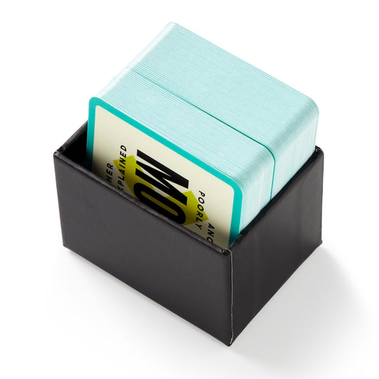 A black box holds Brass Monkey’s Mini Poorly Explained Movies - 80s Edition, a stack of turquoise-edged cards ideal for party games, with the top card showing bold black text on a yellow and white background.