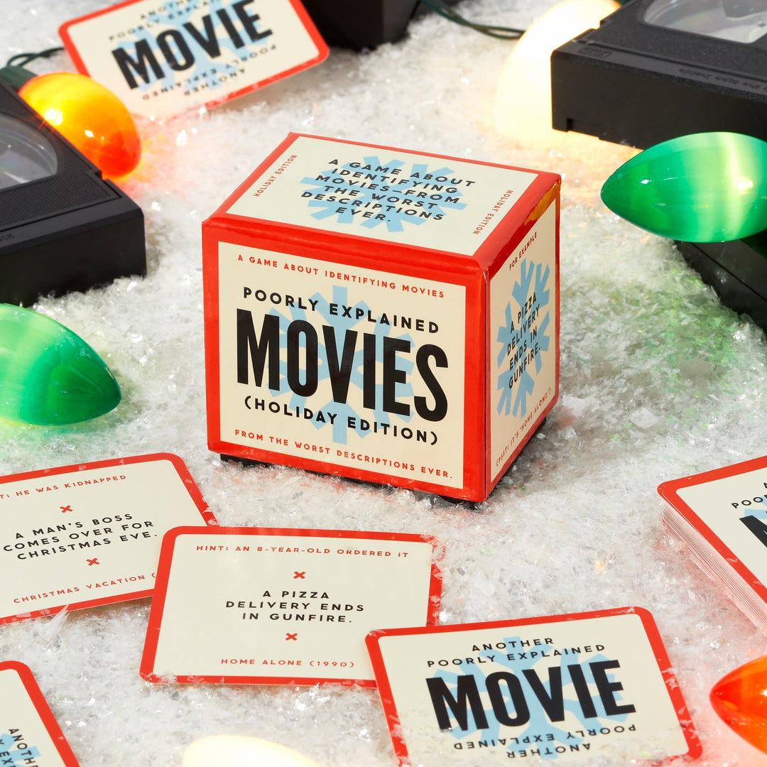 A festive tabletop with snow, Christmas lights, VHS tapes, and red-bordered cards surrounds a cube box labeled “Mini Poorly Explained Movies - Holiday Edition” by Brass Monkey—a party game of holiday movies and wild summaries.