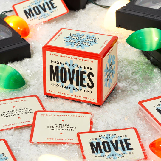 A festive tabletop with snow, Christmas lights, VHS tapes, and red-bordered cards surrounds a cube box labeled “Mini Poorly Explained Movies - Holiday Edition” by Brass Monkey—a party game of holiday movies and wild summaries.
