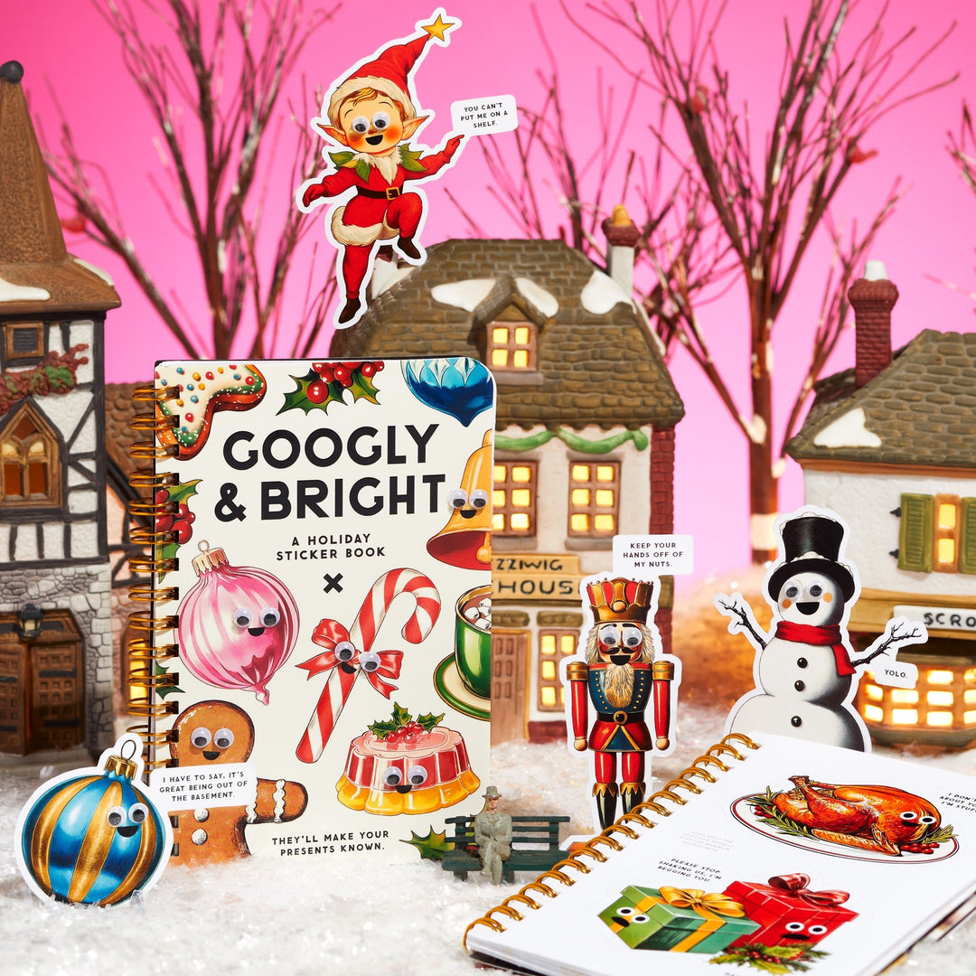 A festive scene features the Brass Monkey Googly & Bright Holiday Sticker Book with stickers of ornaments, an elf, snowman, and nutcracker. Oversized stickers are displayed among model houses and snowy decor on a pink backdrop with bare trees.