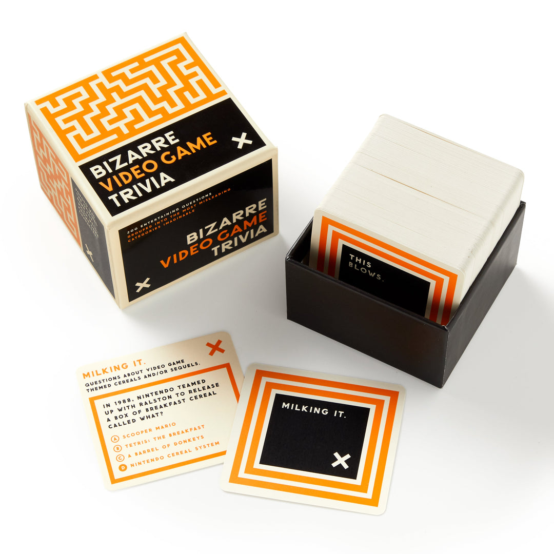 A box of Brass Monkey’s Bizarre Video Game Trivia is open, revealing a stack of question cards in a black holder. Two cards are displayed—one shows quirky video game trivia and the other reveals the answer, Milking it.