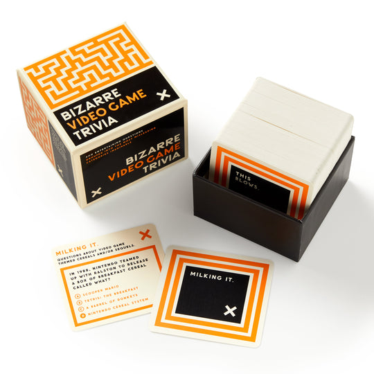 A box of Brass Monkey’s Bizarre Video Game Trivia is open, revealing a stack of question cards in a black holder. Two cards are displayed—one shows quirky video game trivia and the other reveals the answer, Milking it.