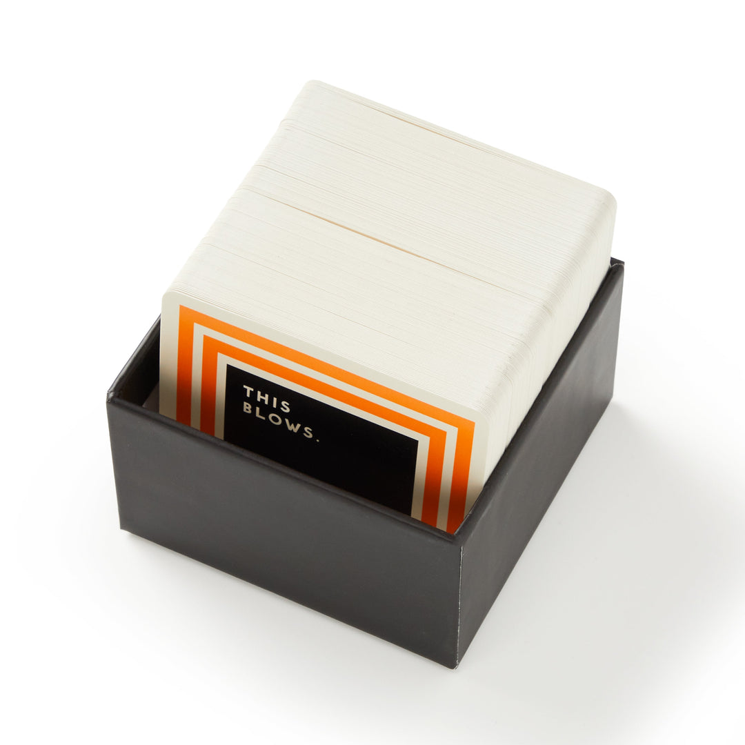 A black box contains a stack of playing cards with white backs; the top card has an orange border and says THIS BLOWS. in white on black—perfect for fans of quirky facts or Bizarre Video Game Trivia by Brass Monkey.