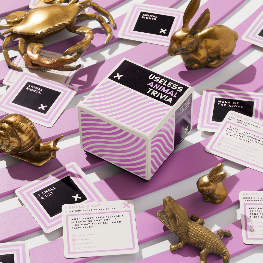 The Brass Monkey Useless Animal Trivia cube rests on a purple and white striped surface, surrounded by animal figurines and trivia cards—inviting you to explore quirky, hidden animal facts.
