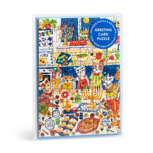 The Happy Hanukcats Greeting Card Puzzle by Anna Ivanir features festive cats celebrating Hanukkah with a menorah, food, and decorations. Packaged with a blue “Greeting Card Puzzle” label.