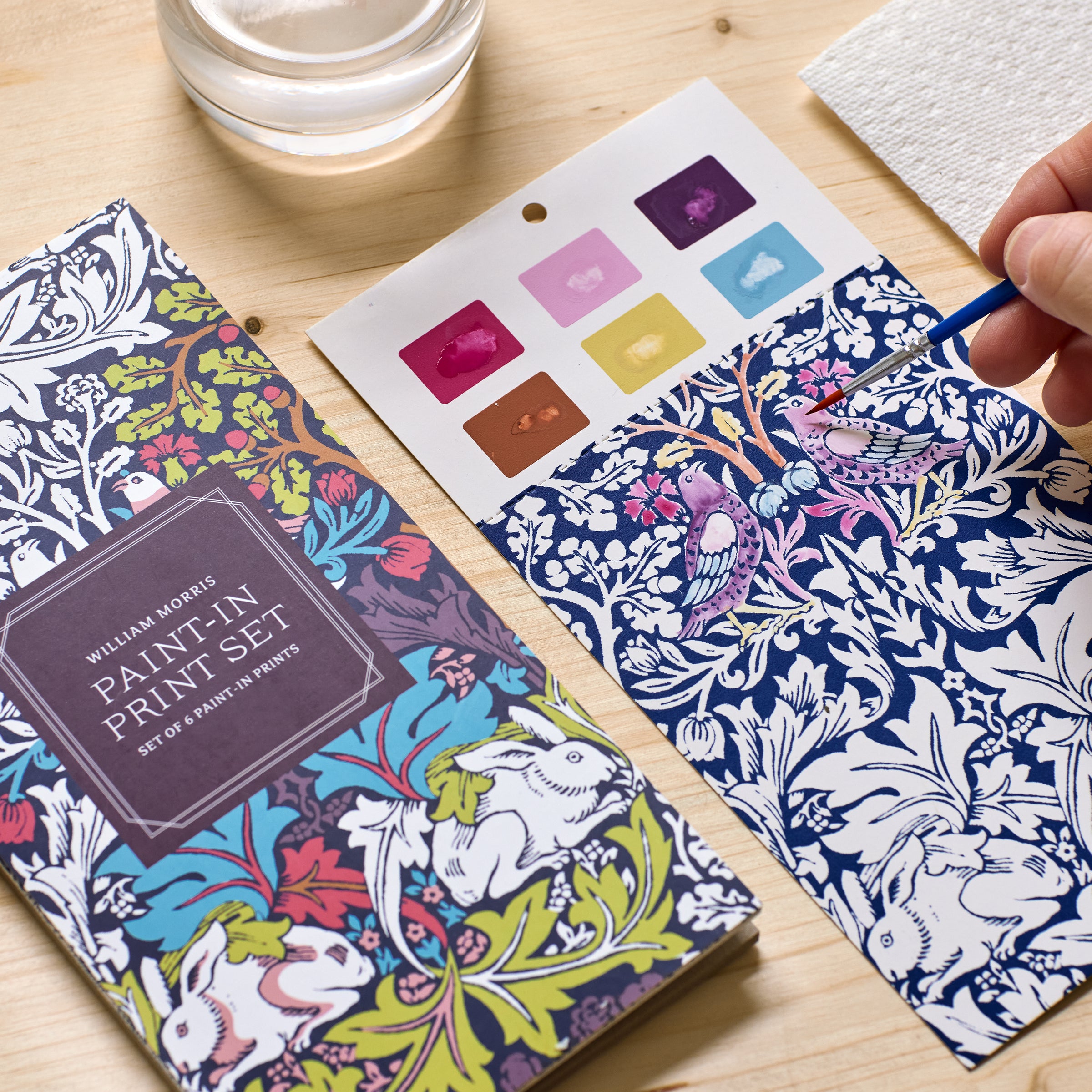 A hand uses a paintbrush and watercolors to fill in a floral and bird design on a coloring sheet from the William Morris Paint-In Print Set. A color swatch card, water cup, and paper towel are also on the table.